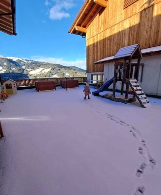 The snowy outdoor area of the Bauernhof, featuring a children's playground and mountain views.