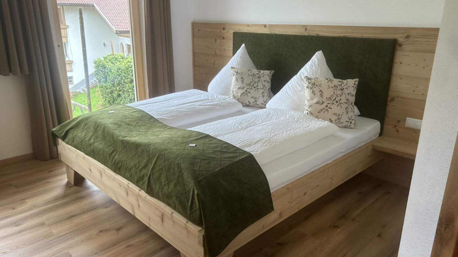 Double bedroom in the farm house with a wooden bed frame, green upholstered headboard, and a window view of residential buildings and greenery.
