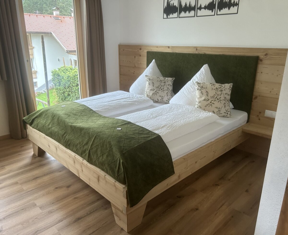 Double bedroom in the farm house with a wooden bed frame, green upholstered headboard, and a window view of residential buildings and greenery.