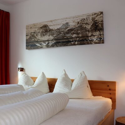 A cozy bedroom in the apartment featuring a wooden bed with plush white bedding, rustic mountain wall art, and warm reading lights.