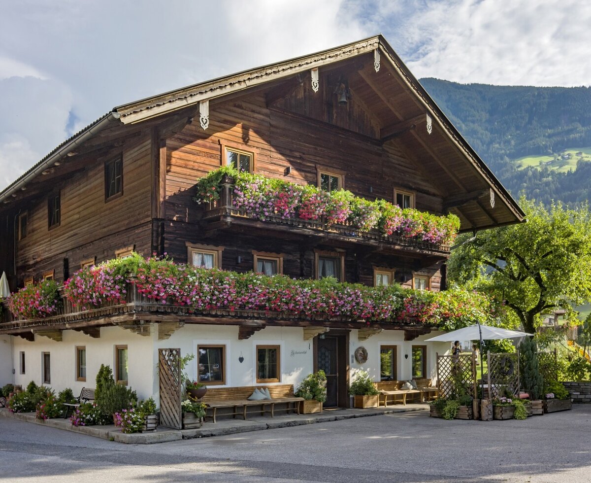 The Farm House exterior features traditional wooden construction, balconies with flowers, outdoor benches, and a view of the mountains.