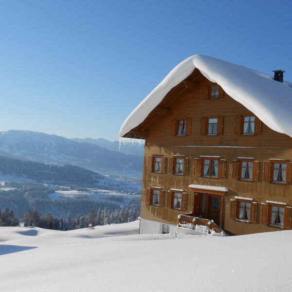 The Farm House exterior in winter, covered in snow, with icicles hanging from the roof and a mountain backdrop.
