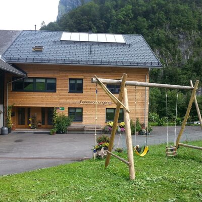 The Farm House exterior features a wooden facade, solar panels on the roof, and a swing set on the lawn in front of the entrance.