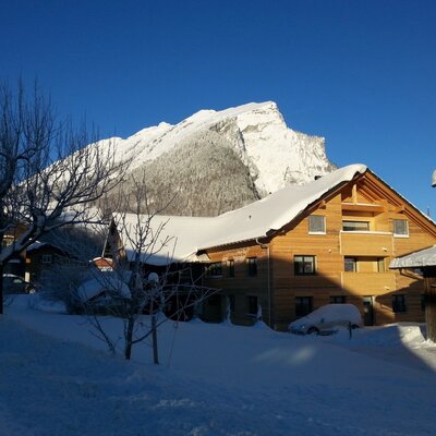 The wooden farm house exterior during winter, with snow on the roof and ground, and a snow-covered mountain visible behind it.