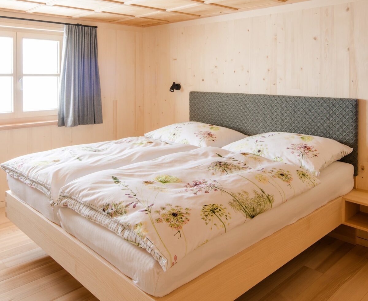 Double bed with floral bedding in a Farm House bedroom, featuring wooden paneled walls, a window, and an integrated bedside table with an electrical outlet.