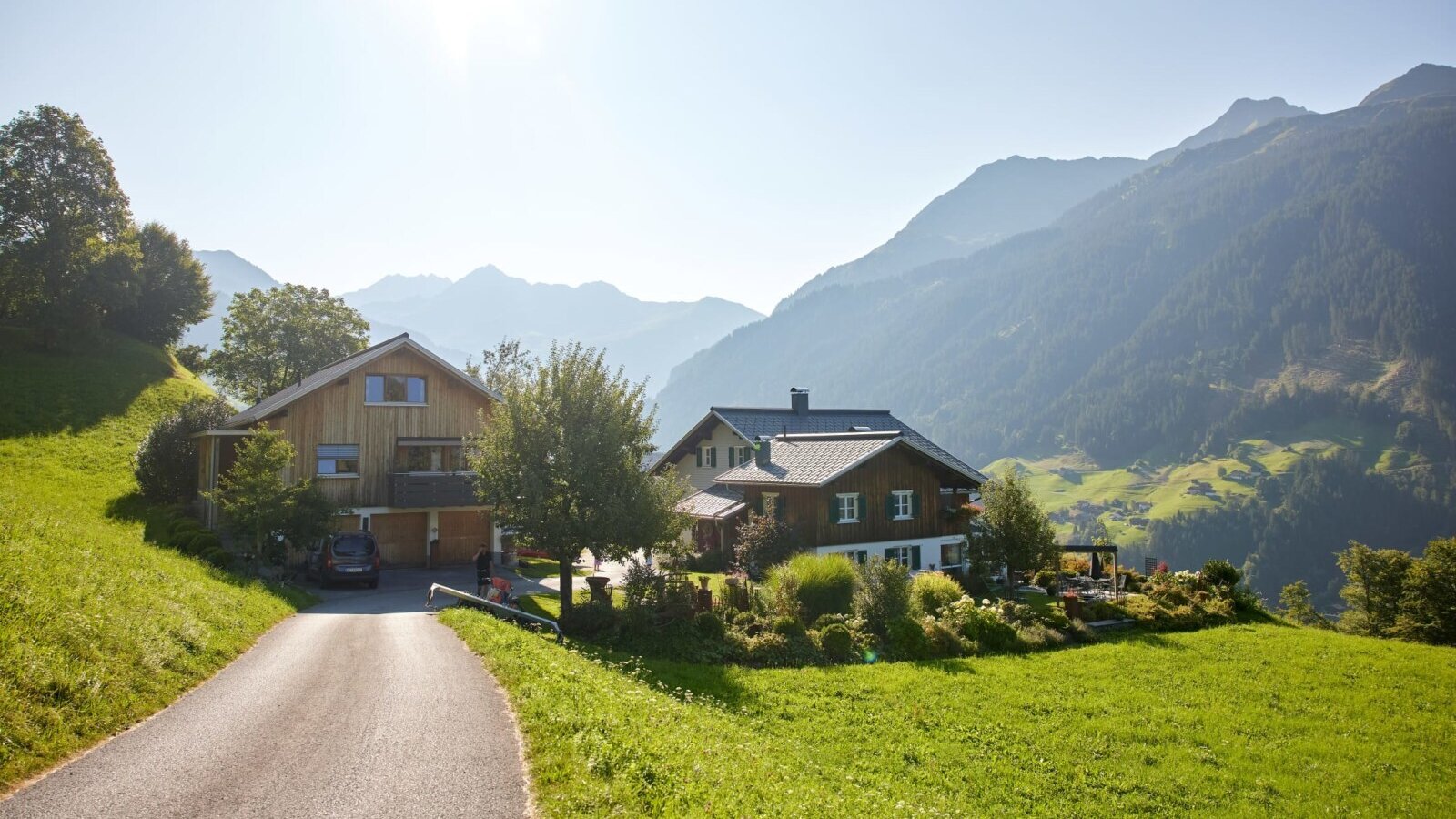Exterior view of the farmhouse with an access road, surrounded by green meadows and the Alps in the background.