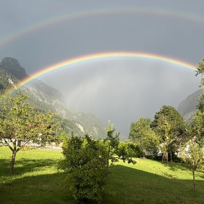 Double rainbow over the mountains, visible from the farmhouse with a green lawn and trees.
