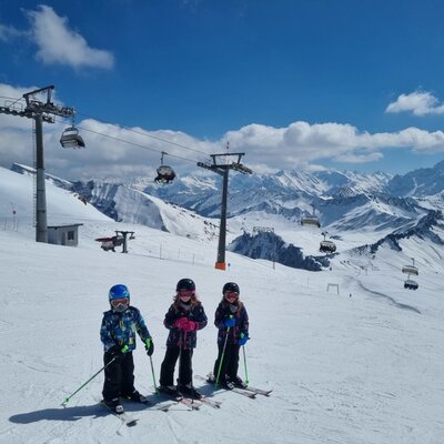 Three children on a ski slope with ski lifts and snow-covered mountains in the background.