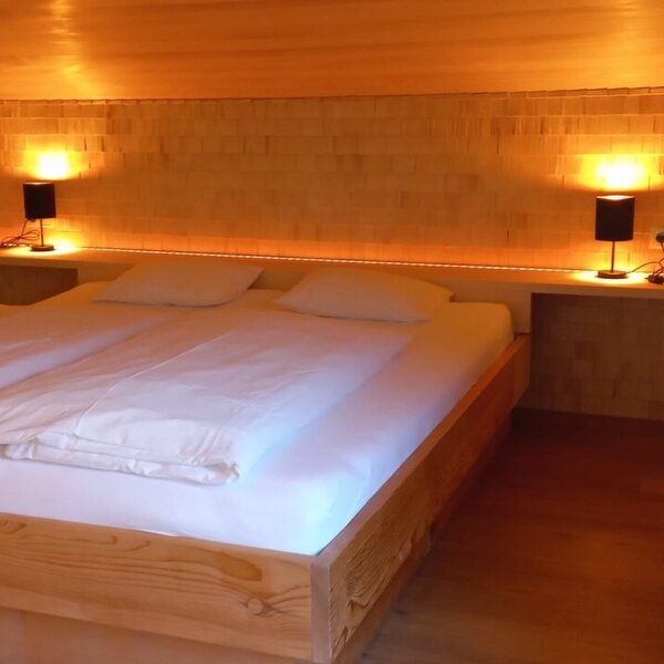 Double bed in the bedroom of the farmhouse with wooden walls, integrated bedside tables, and reading lamps.