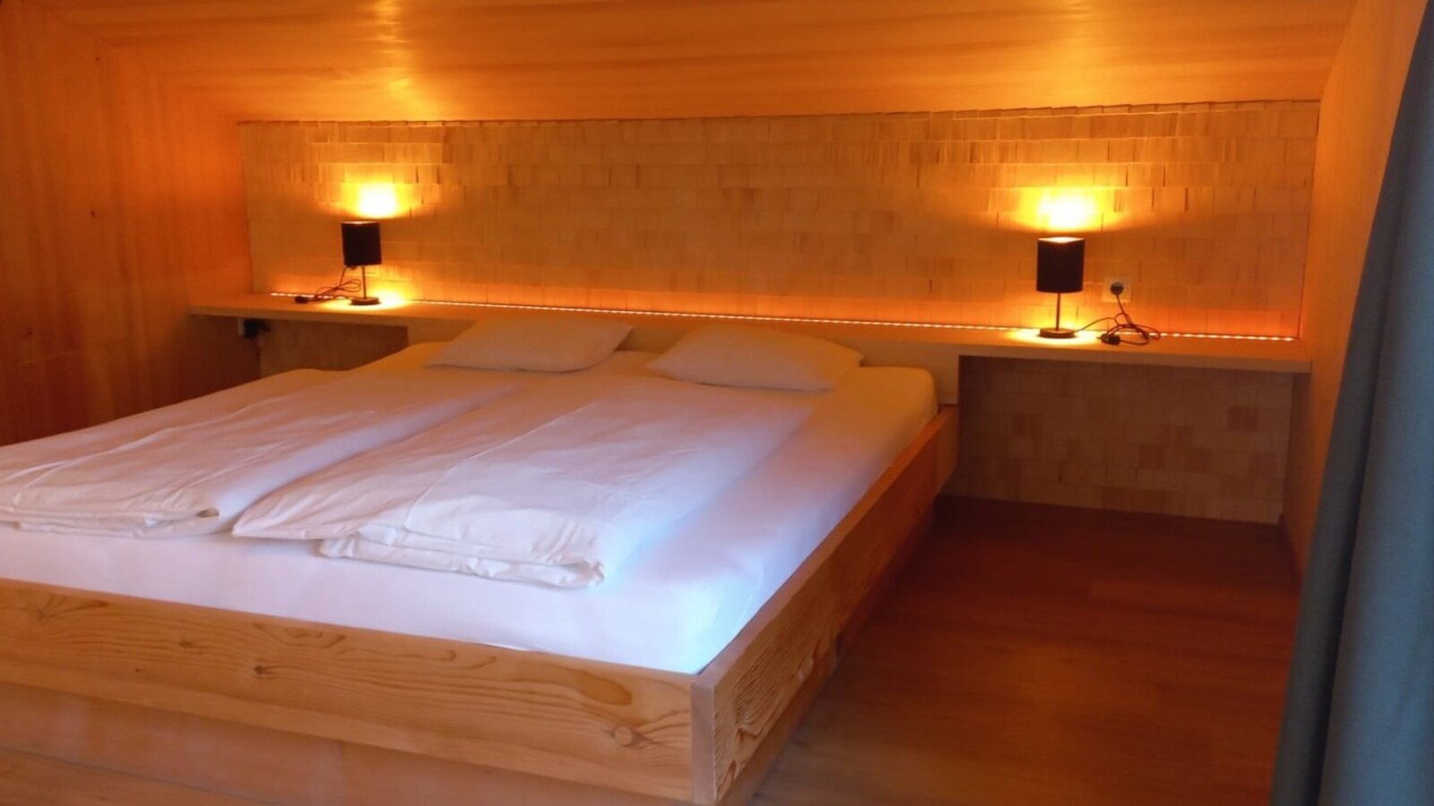 Double bed in the bedroom of the farmhouse with wooden walls, integrated bedside tables, and reading lamps.