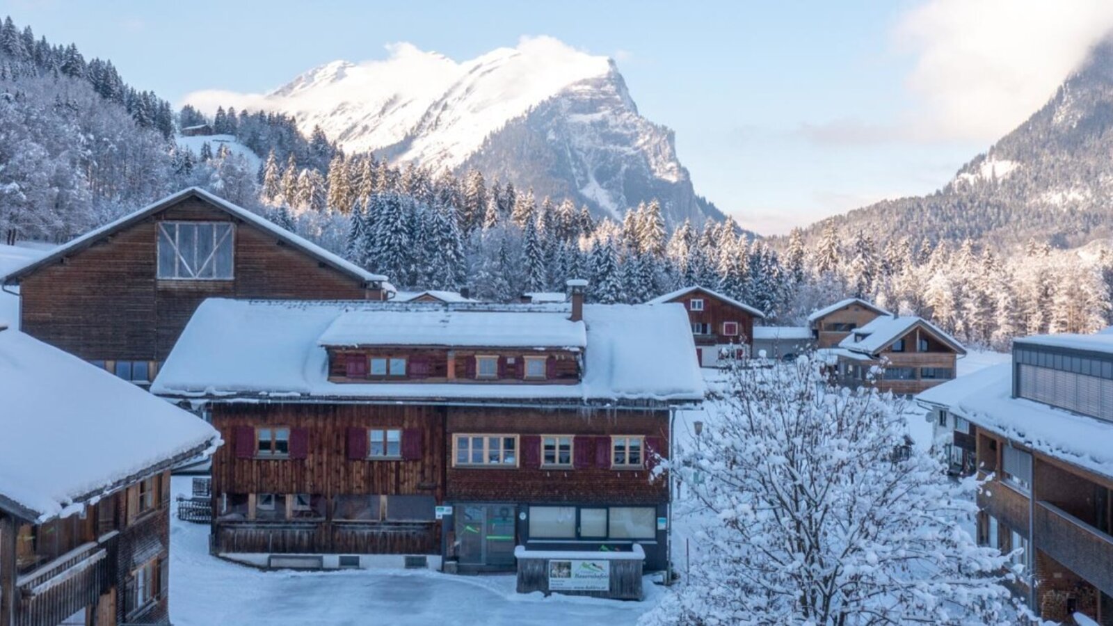 The Farm House exterior and surrounding buildings are covered in snow, set against a backdrop of snowy mountains.