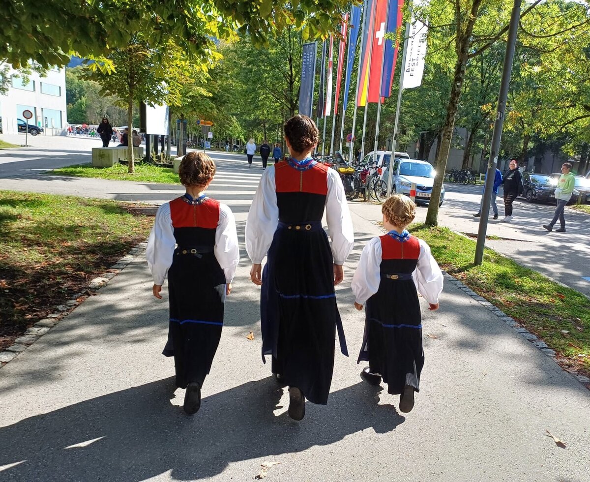 Three people in traditional attire walking on a paved path lined with trees and flags.