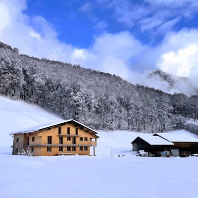 The holiday farm with a wooden facade and balconies, set in a snow-covered winter landscape with forests and mountains.