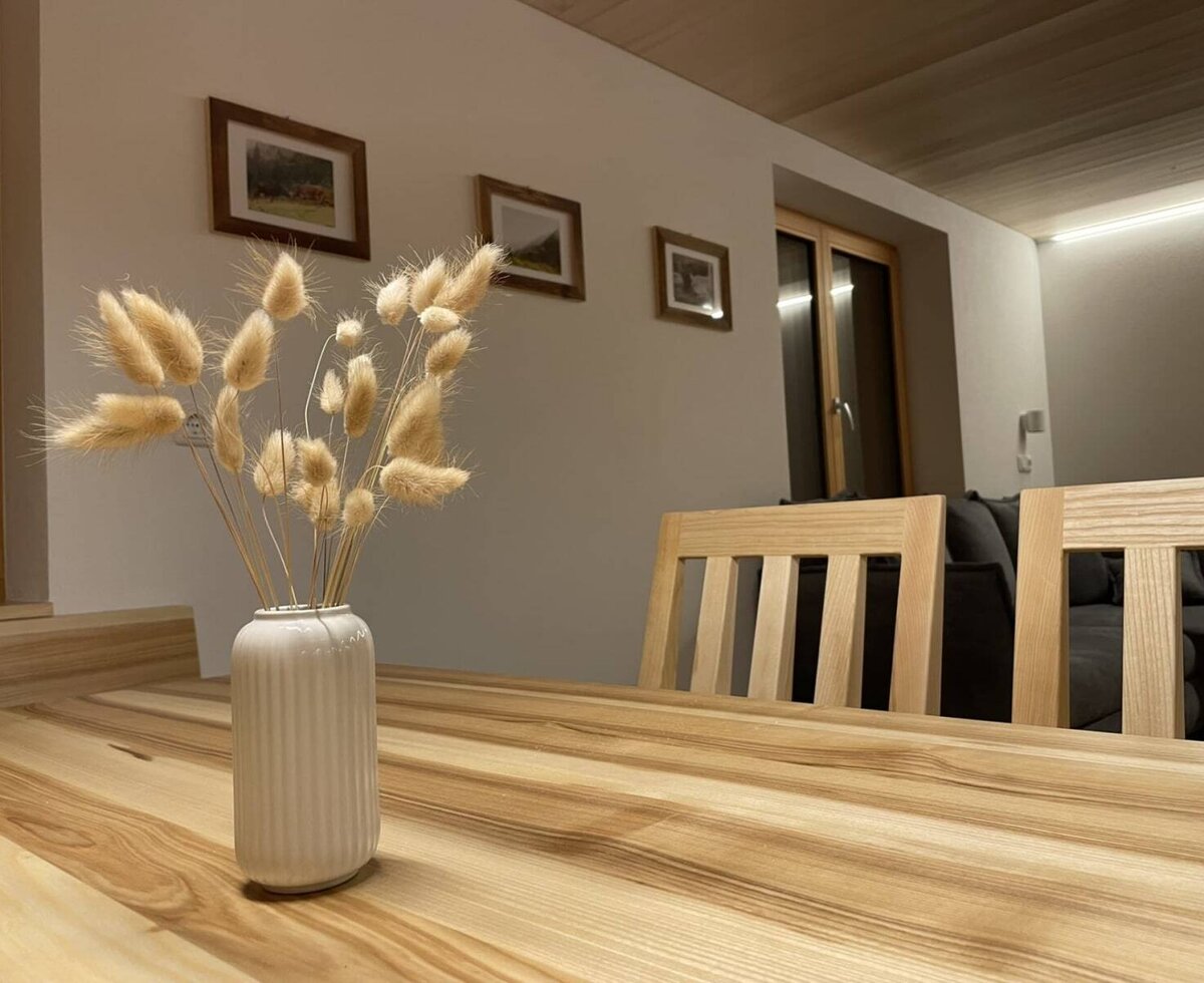Wooden dining table in the appartment with a vase, chairs, and a sofa in the background.
