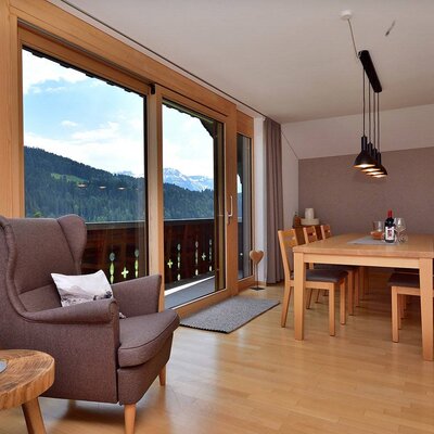 The farm house features an open-plan living and dining area with a wooden table, chairs, a comfortable armchair, and sliding glass doors that open to a balcony with mountain and forest views.