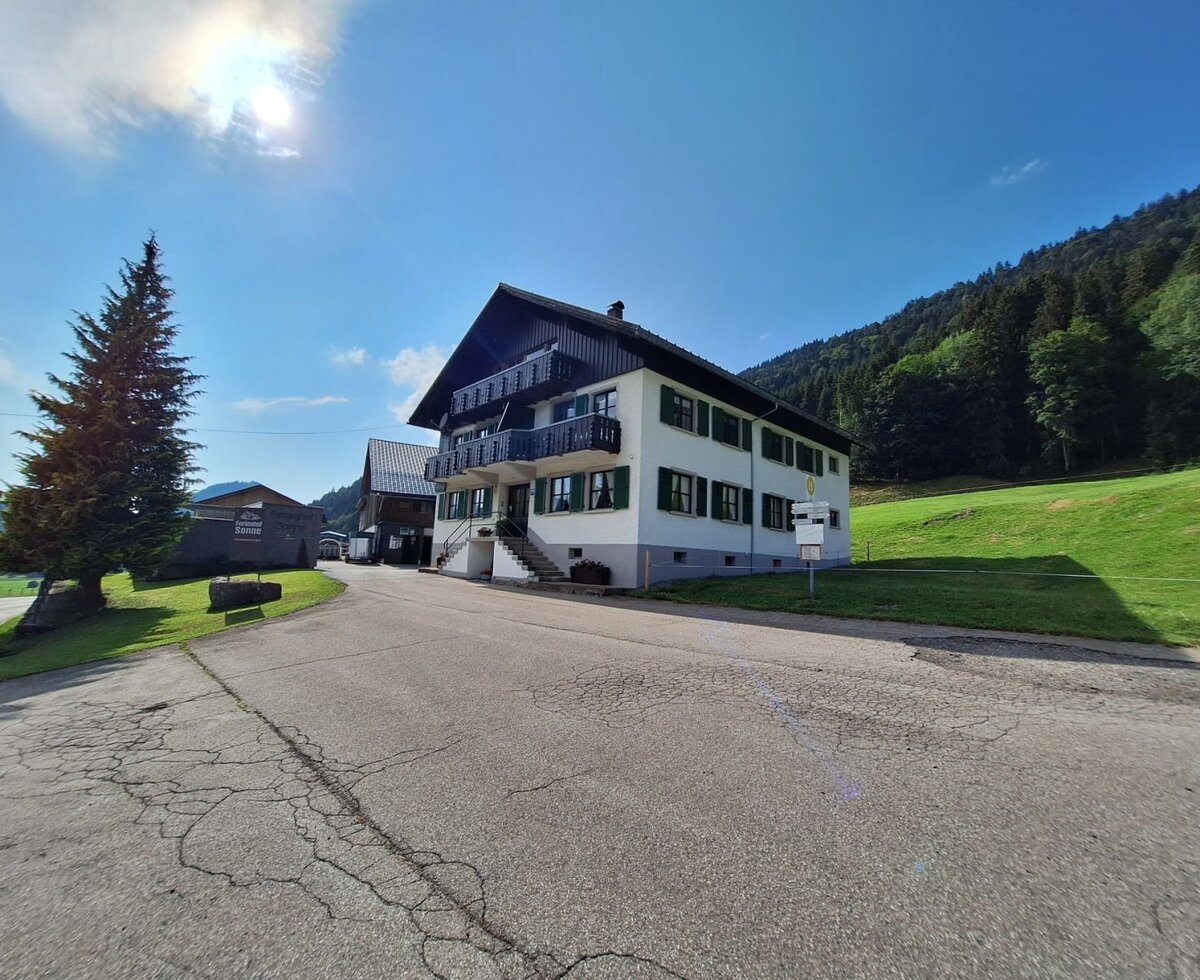 The inviting farmhouse under a bright blue sky, surrounded by green meadows and forested slopes.