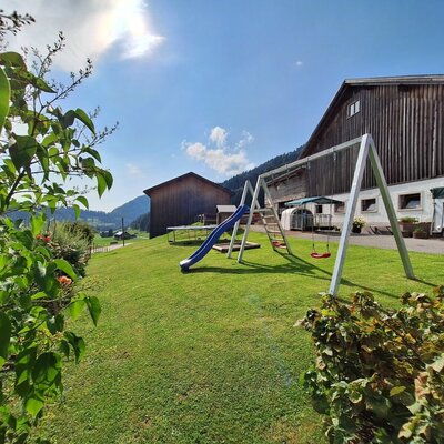 The farmhouse's inviting garden featuring a sunny lawn, a children's playground, and scenic mountain views.