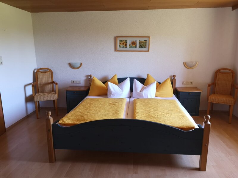 Farm House bedroom featuring a double bed with yellow bedding, two bedside tables with wall lights, and two wicker armchairs.