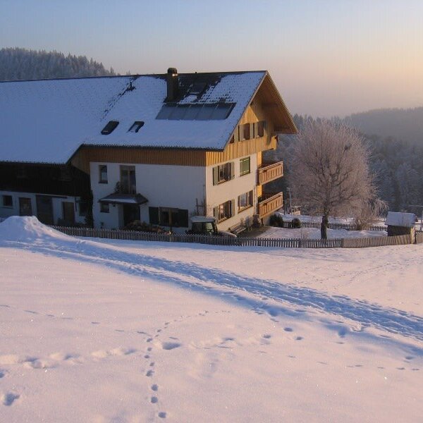 The Farm House exterior in winter, featuring a snow-covered roof, balconies, solar panels, and a fenced yard surrounded by snowy fields.