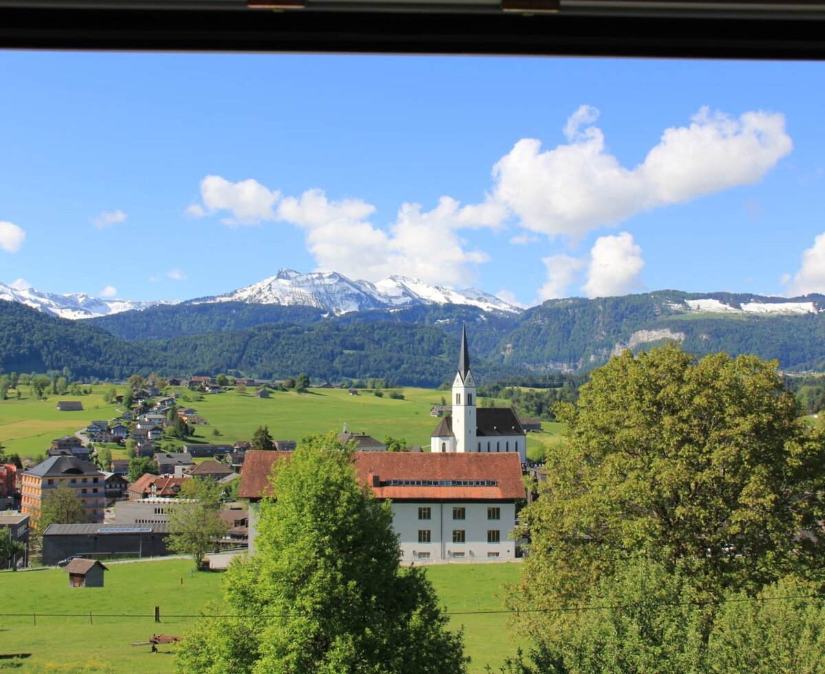 View from the farmhouse window of the village with a church, green fields, and snow-capped mountains.