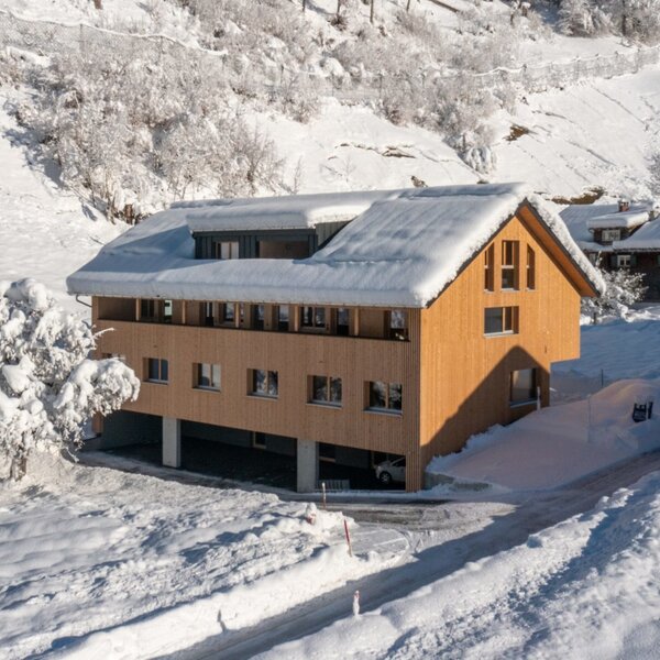 The modern wooden farmhouse in a snow-covered mountain setting, featuring covered parking beneath the building.