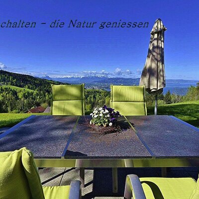 An outdoor seating area at the Farm House features a table and chairs with views of green hills, a lake, and distant mountains.