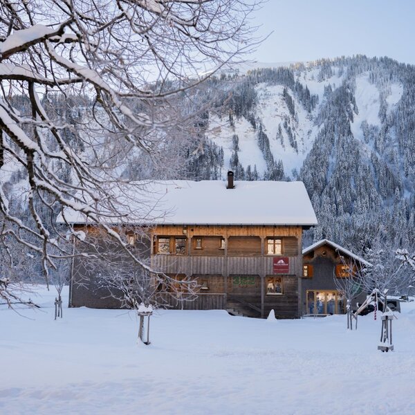 The Farm House exterior, a wooden building covered in snow, with illuminated windows against a backdrop of snow-covered mountains. The Farm House exterior, a wooden building covered in snow, with illuminated windows against a backdrop of snow-covered mountains.