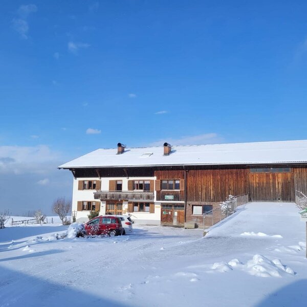 The farmhouse in winter, with a snow-covered roof, white facade, and an adjacent wooden barn under a clear blue sky.
