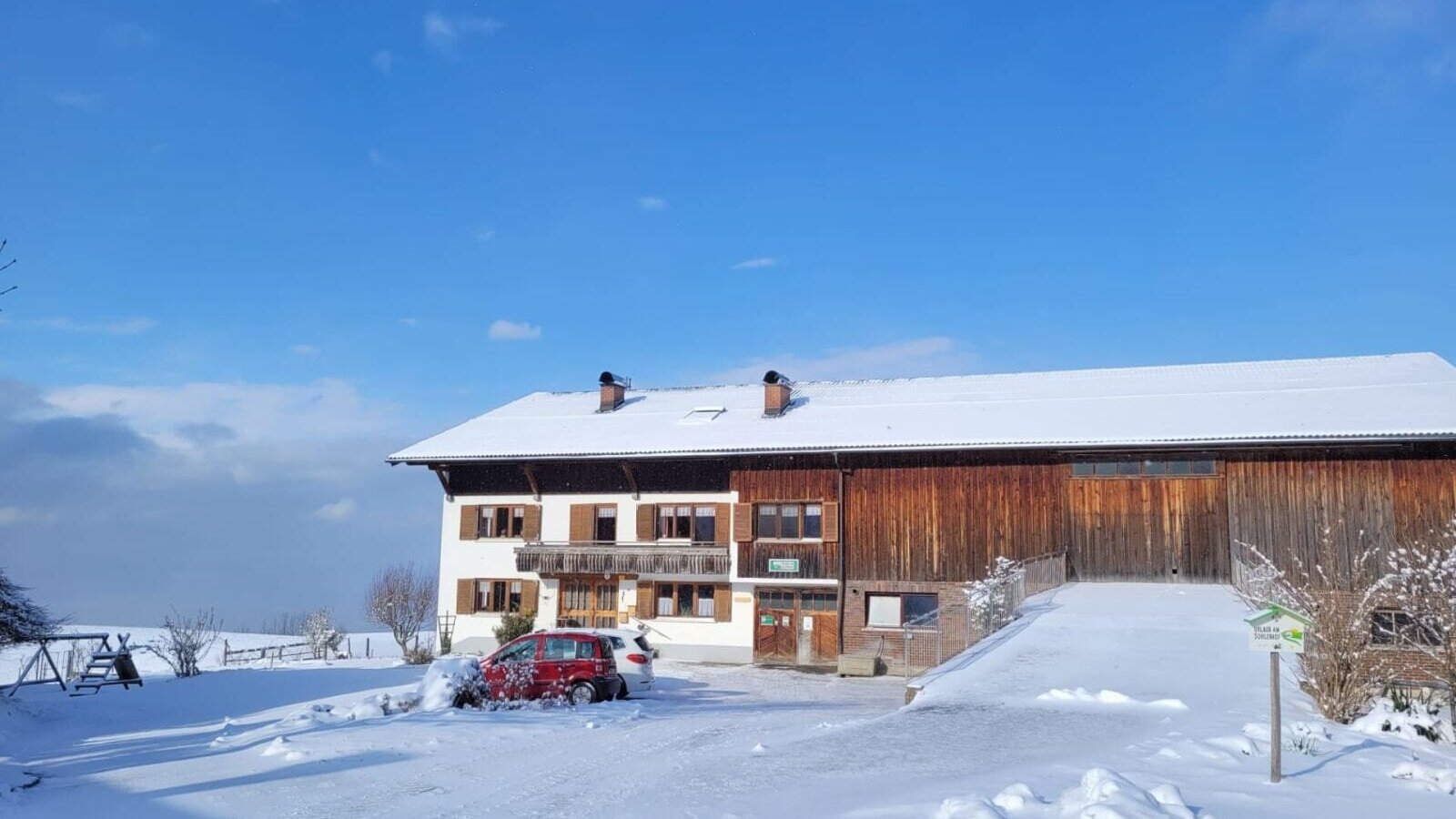 The farmhouse in winter, with a snow-covered roof, white facade, and an adjacent wooden barn under a clear blue sky.