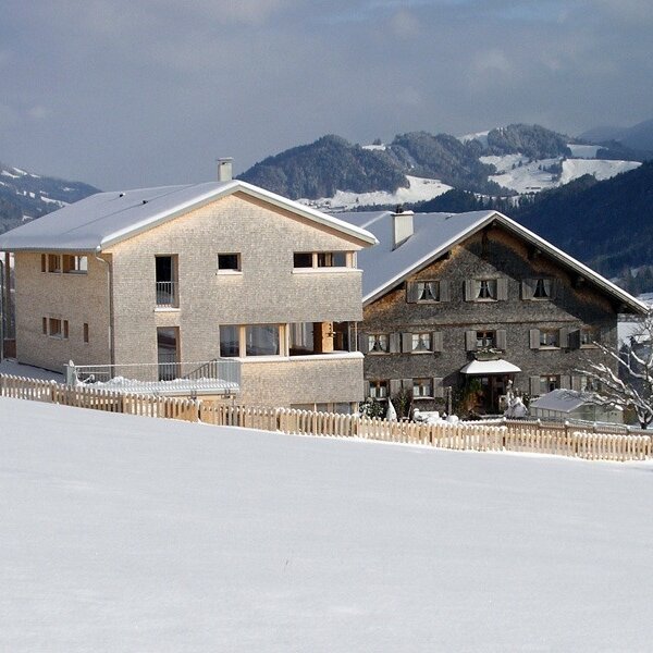 The two farm houses, a modern light-wood structure and a traditional dark-wood building, surrounded by snow-covered fields and mountains. The two farm houses, a modern light-wood structure and a traditional dark-wood building, surrounded by snow-covered fields and mountains.