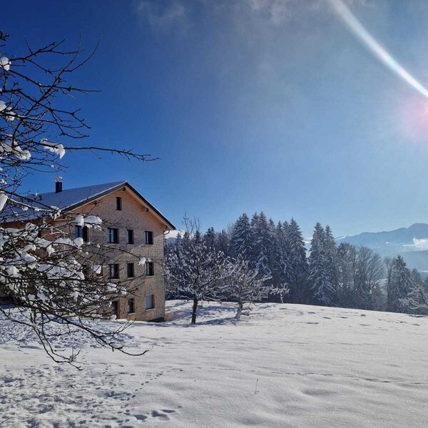 The farmhouse in a snowy winter landscape with trees and mountains under a blue sky.