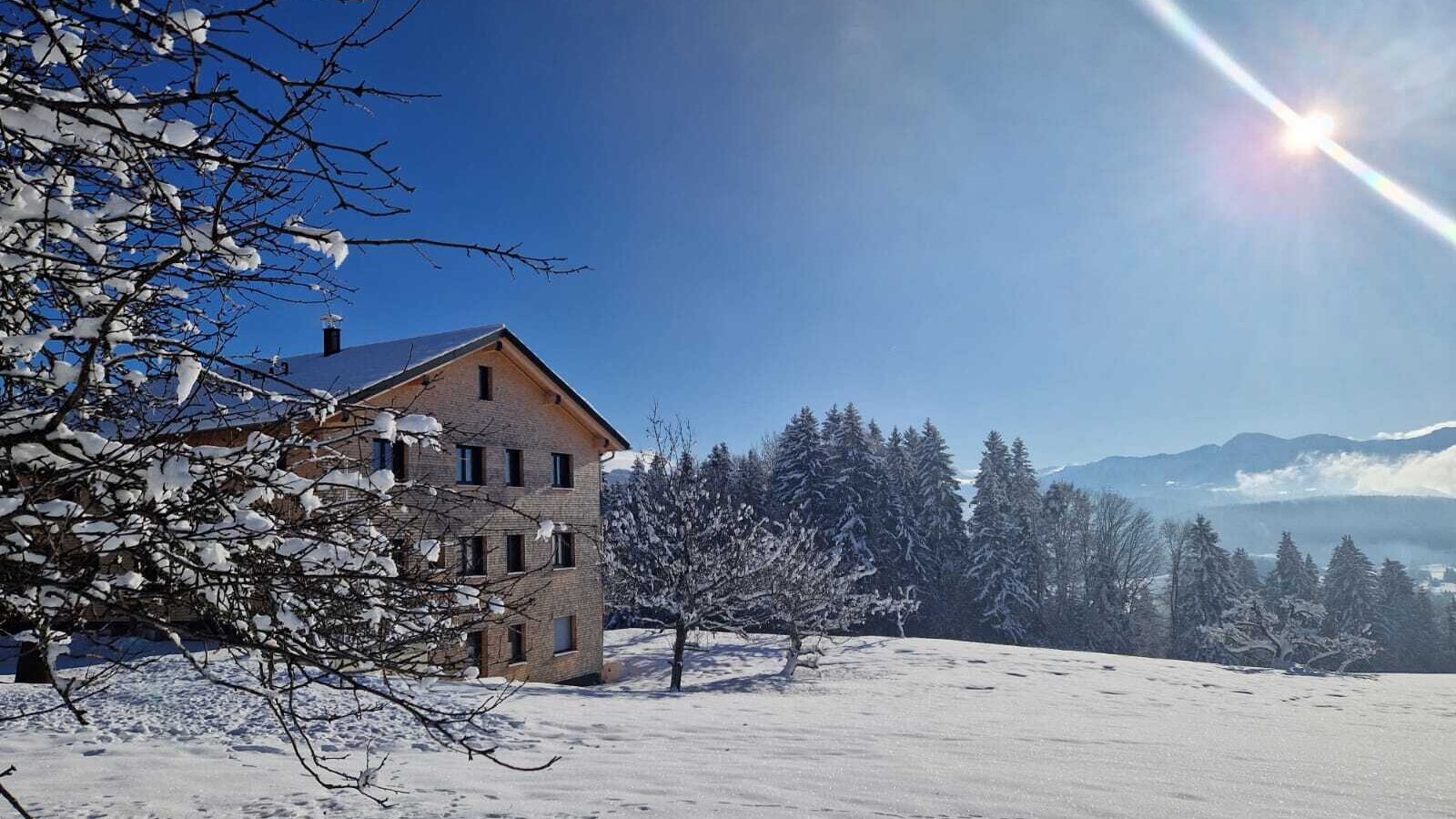 The farmhouse in a snowy winter landscape with trees and mountains under a blue sky.