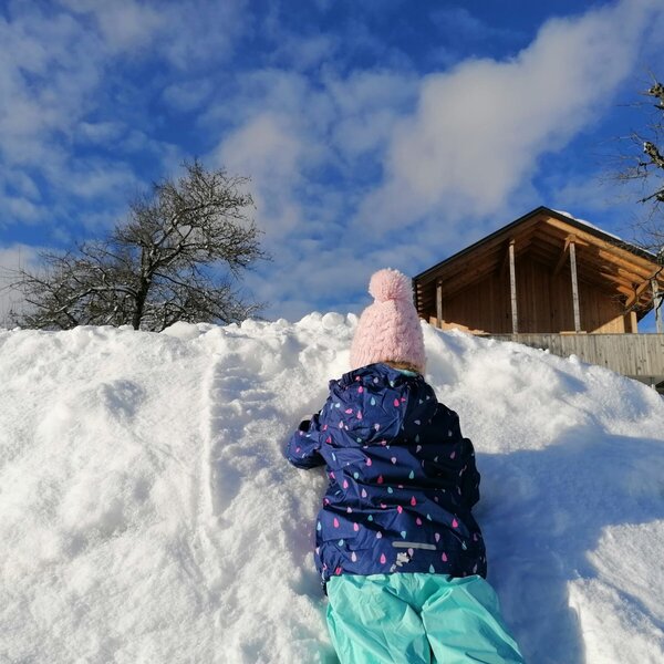 A child plays in the snow on the Farm House grounds, with the wooden building visible in the background.